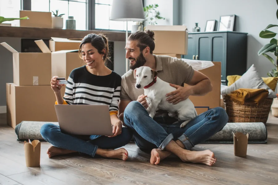 Couple and dog in new home, reviewing finances on laptop for house savings.