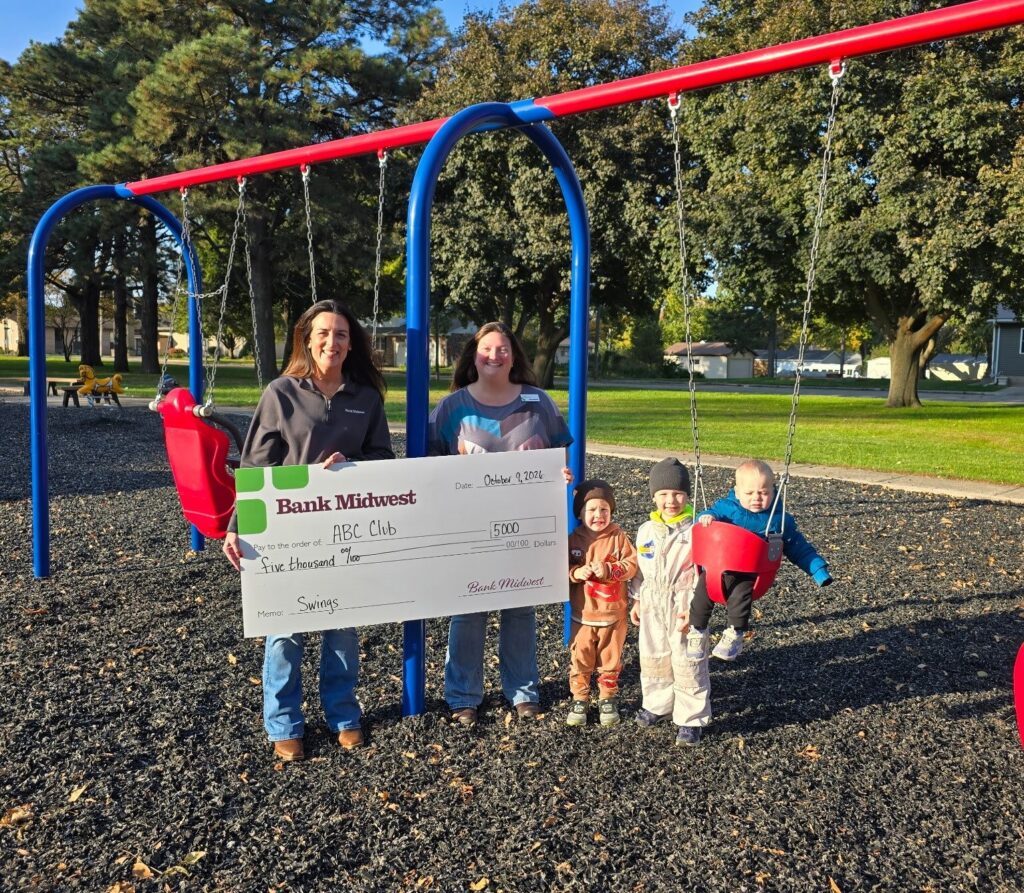 Image of check presentation from Bank Midwest to Armstrong Betterment Club. Photo is taken at the city park with kids in the swing. 